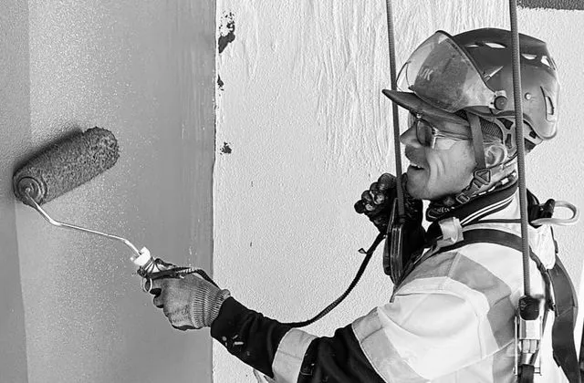 Alpine-Tech 360 rope access technician painting a building wall with a roller while suspended on ropes